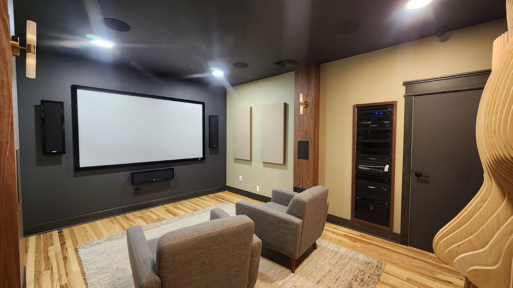 A media room with a projection screen, fabric acoustic panels, and a sculpted wood accent wall, with an equipment rack visible through a side doorway.